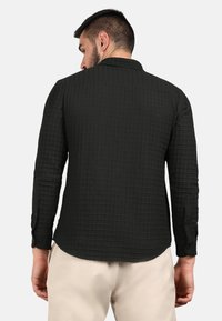Camicia nera a maniche lunghe, strutturata, con un motivo a griglia e polsini abbottonati, caratterizzata da un orlo leggermente curvo e un colletto.