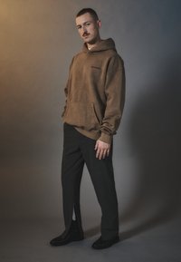 Sudadera oversized marrón con un bolsillo frontal y un logo, combinada con pantalones negros ajustados que tienen una abertura lateral. Llevado con zapatos negros.