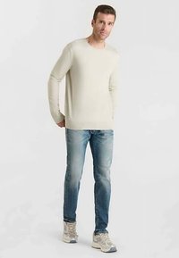 Hellbeiger Langarm-Pullover, eng geschnittene blaue Jeans und hellgraue Sportschuhe mit dunklen Akzenten, präsentiert an einem männlichen Modell.