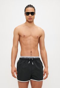 Pantaloni de baie negri cu margini albe și un cordon elastic etichetat "COPENHAGEN STUDIOS." Modelul poartă ochelari de soare, are torsul gol și adoptă o poziție neutră.