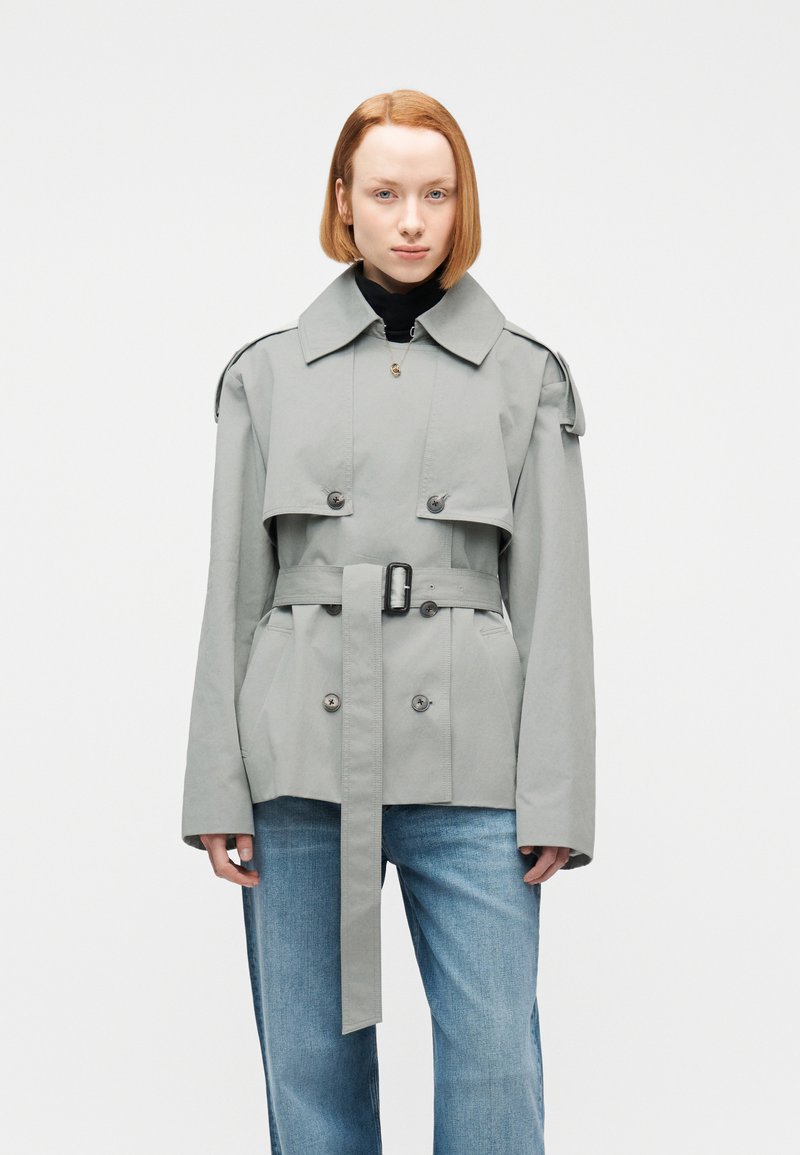 Γυναίκα με κοντά κόκκινα μαλλιά, ντυμένη με γκρι παλτό βραδύκοκκο με ζώνη πάνω από μαύρο μπλουζάκι και μπλε τζιν, στέκεται μπροστά σε απλό λευκό φόντο.