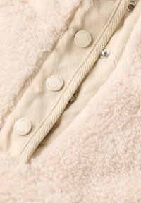 Cream-colored fleece fabric with textured surface, featuring a beige button placket with embossed brand logo and thin striped accents.