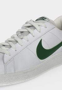 Λευκό αθλητικό παπούτσι Nike με πράσινο λογότυπο swoosh, λευκά κορδόνια και υφασμένο πέλμα σε κοντινή γωνία.