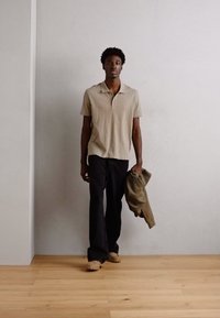 Polo shirt beige con un motivo testurizzato, pantaloni larghi neri e una giacca grigia adagiata su un braccio. Pavimento in legno chiaro e sfondo semplice.