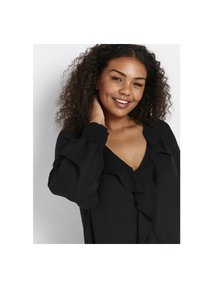 Leende kvinna med lockigt hår iklädd en svart blus med volang vid V-halsen och elastiska manschetter, som rör vid sin hals mot en neutral bakgrund.
