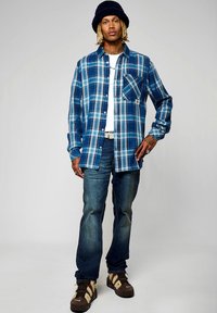 Camisa de cuadros azules con un diseño de botonadura, jeans de mezclilla y zapatillas oscuras. Incluye un bolsillo en el pecho y se superpone con una camiseta blanca.