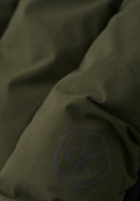 Olivgrön jacka i tyg med en slät textur, med en svart logotyp framtill. Designen är strömlinjeformad med minimala detaljer.