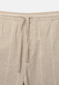 Pantalon en maille beige avec taille élastique et cordon noué au centre devant, présentant des détails de coutures verticales.