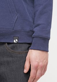Felpa blu navy con tessuto strutturato e polsini a coste, dotata di un'etichetta nera con il logo vicino all'orlo, abbinata a jeans scuri.