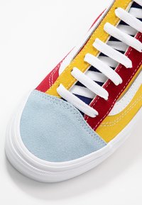 Baskets multicolores avec des sections en toile rouge, jaune et bleue, un capuchon de chaussure en suède bleu doux, et des lacets blancs avec des coutures contrastées.