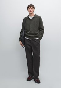 Olijfgroene trui met een v-hals, gedragen over een wit shirt. Donkere broeken en bordeauxrode schoenen. Zwarte tas over de schouder.