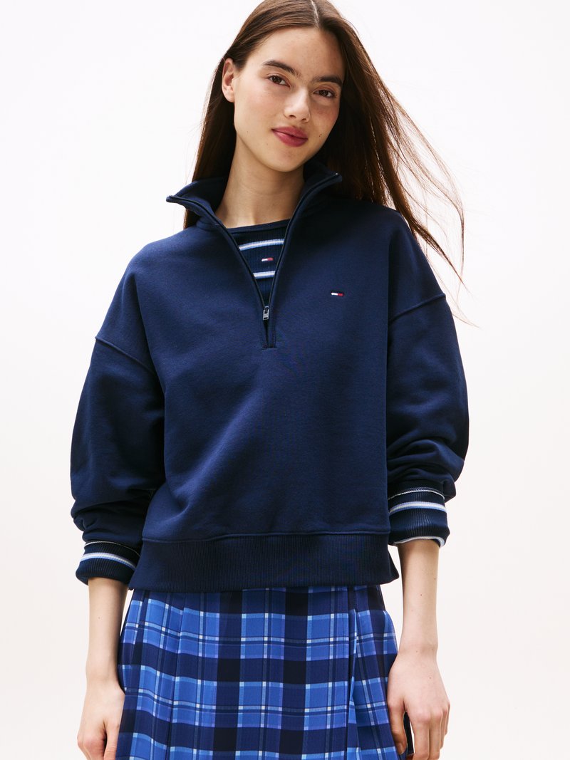 Sweatshirt azul-marinho com fecho zip, colarinho alto, punhos canelados com riscas azuis e brancas, sobre uma saia xadrez azul.