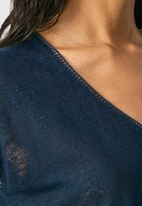 Tmavě modrý pletený top s výstřihem do V, s texturovanou tkaninou a jemným otevřeným švem podél okraje.