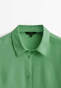Chemise verte à manches courtes en tissu léger avec un col et cinq boutons, présentant une apparence texturée et légèrement froissée.