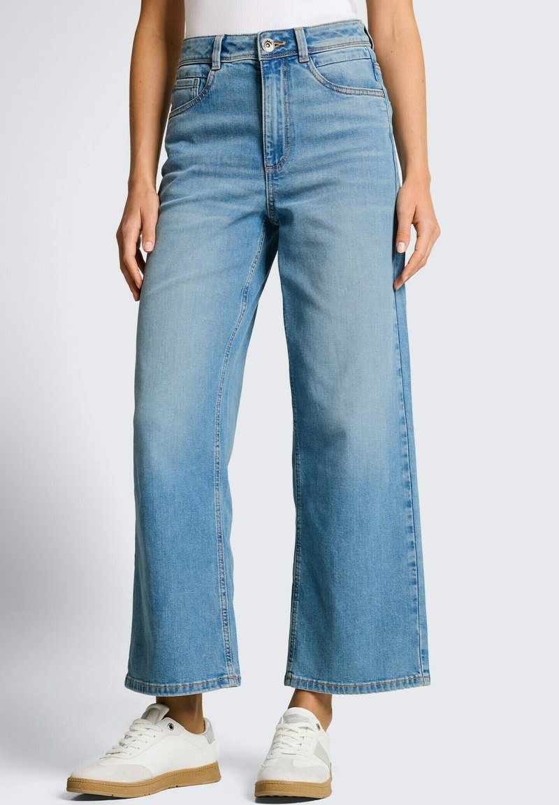 Femme portant un jean taille haute à jambes larges bleu clair associé à des baskets blanches sur un fond uni.