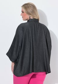 Blonde Frau mit silbernen Creolen, einem dunkelgrauen, lockeren Oberteil mit weiten Ärmeln und leuchtend pinken Hosen, steht vor einer weißen Wand.
