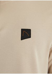 Beige T-Shirt mit einem schwarzen quadratischen Patch, der ein geometrisches oranges Design zeigt. Der Stoff wirkt glatt und dehnbar.