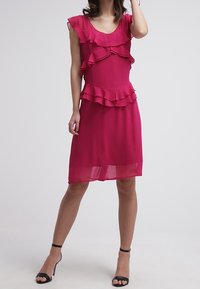 Vestido fucsia con detalles de volantes en capas, mangas cortas y un corte a la altura de la rodilla. Está hecho de una tela ligera, transparente en el dobladillo.