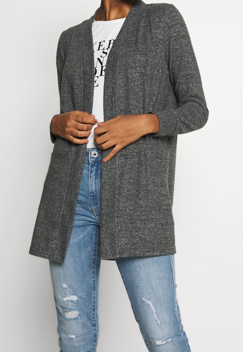 Cardigan largo gris con frente abierto, textura de punto y mangas largas. Combinado con una camiseta blanca y jeans desgastados en azul claro.