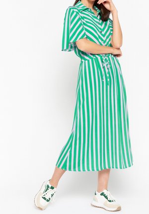 Femme portant une robe midi à rayures verticales vertes et blanches avec des boutons et des baskets, posant avec une main près de son visage.