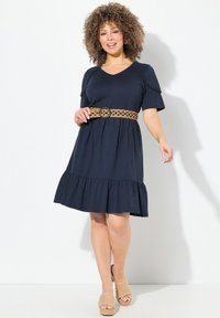 Tummansininen lyhyttä hihaista mekkoa, jossa on röyhelöhelma, on yhdistetty kudottuun beigenväriseen vyöhön. Mallilla on päällään vaaleanruskeat korot sandaalit.