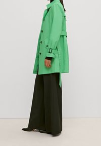 Manteau d trench vert avec un bouton avant, taille ceinturée et accents noirs, associé à un pantalon noir à jambes larges et des chaussures noires pointues.