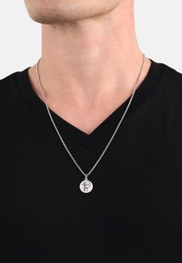 Silverfärgad halskedja med en rund hängande berlock som har en detaljerad design, bärs över en svart v-ringad tröja. Kedjan är delikat och metallisk.
