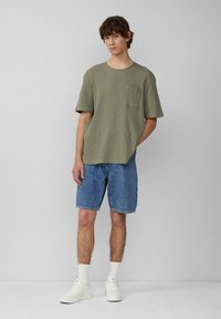 Oliwkowa, fakturowana koszulka z krótkim rękawem z kieszenią na piersi, w połączeniu z dżinsowymi szortami i białymi trampkami, na jednolitym tle.