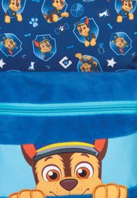 Blauwe tas met een cartoonhond met grote ogen, een glimlach en een pet. De achtergrond heeft kleurrijke pootafdrukken en badges. Met een ritsluiting.