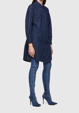Femme portant une longue chemise en denim bleu foncé avec des poches à l'avant et des bottes hautes en denim à talons assorties.