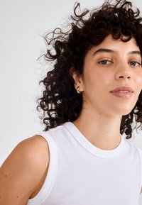 Hvid ærmeløs top med rund halsudskæring, lavet af glat stof. Vist med krøllet hår, gule hoop øreringe og subtil makeup.