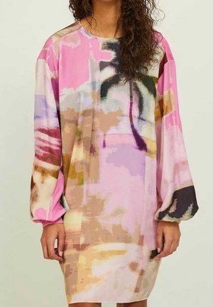 Vrouw draagt een loszittende roze jurk met lange mouwen en abstracte meerkleurige patronen, staand tegen een effen lichte achtergrond.