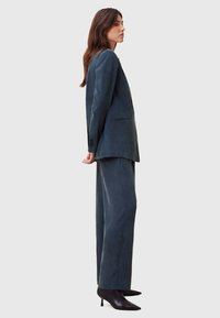 Maatwerk donkergroen pak van gladde stof; beschikt over een enkelvoudig jasje en brede pantalons, aangevuld met zwart puntige hakken.