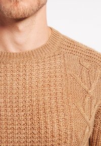 Maglione lavorato a maglia grossa in marrone chiaro, con motivi a coste e a trecce. La vista ravvicinata mette in risalto il filato testurizzato e il collo a giro.
