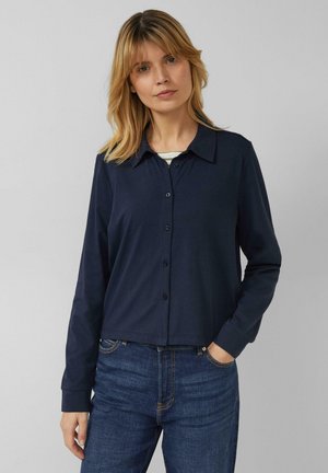 Vrouw met schouderlang blond haar, gekleed in een marineblauw overhemd met knopen en lange mouwen, en een blauwe spijkerbroek, staand met één hand in haar zak.