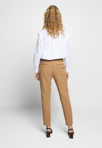 Kraftigt skräddarsydda byxor i tan med smal passform, matchat med en beskuren vit skjorta och svarta klacksandaler. Slätt tyg med subtila detaljer på bakfickorna.