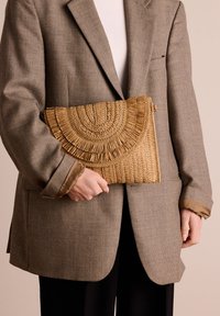 Natuurlijk geweven riet clutchtas met een gebogen franjeslaag, gehouden tegen een gestructureerde grijze blazer, met een lichtbruin kleur.
