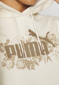 Krämfärgad hoodie med en stor brun "PUMA"-logotyp, florala grafiska detaljer och snoddar vid halslinningen. Mjuk, strukturerad tyg.