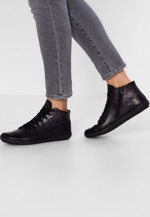 Baskets montantes en cuir noir avec fermeture éclair latérale, portées avec un jean gris raccourci, montrées sur une personne marchant sur fond blanc.