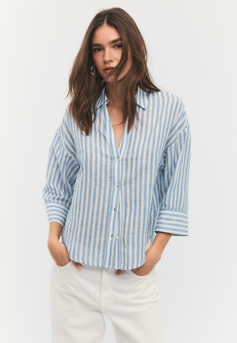 Young woman with loose brown hair wears a blue and white vertical striped button-up shirt and white pants, standing with hands in pockets.