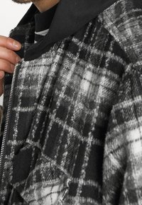 Personne portant une veste à carreaux noir et blanc avec une fermeture éclair, tenant le col près du cou par-dessus un sweat à capuche noir et une chemise rayée.