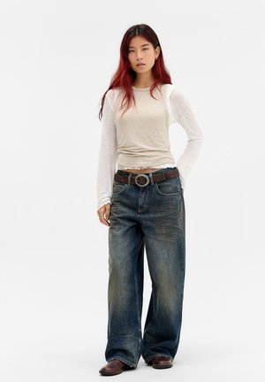 Giovane donna con lunghi capelli rossi indossa maniche lunghe beige e bianche a strati, jeans larghi sbiaditi con cintura marrone e scarpe marroni, in piedi su sfondo bianco.
