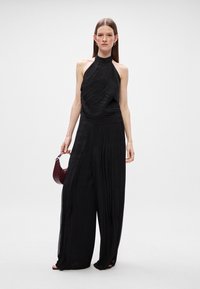 Μαύρο ζιπ-κιλό halter neck με πτυχωτή υφή, σχέδιο φαρδιάς παντελόνι, και διακριτική γυαλάδα; συνδυασμένο με μια μπορντό τσάντα υφής.