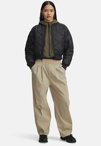 Sort quiltet bomberjakke over en olivengrøn zip-up hoodie, parret med beige plisserede bukser og sorte sneakers.