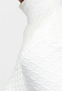 Biała teksturowana odzież z falistym wzorem, z wysokim kołnierzem i widocznymi detalami szewnymi. Materiał wydaje się gładki i elastyczny.