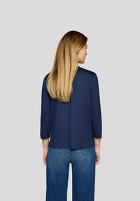 Marineblaue Dreiviertelarm-Bluse mit lässigem Schnitt, glatter Textur und leicht längerem Rückenbereich, kombiniert mit hellblauen Jeans.