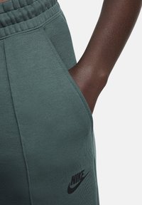 Groene sweatpants van een gestructureerde stof, met een elastische tailleband, zijzakken en een zwart Nike-logo op de linkerdij.