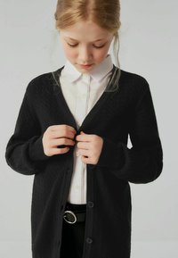 Cardigan nero in maglia con scollo a V, motivo testurizzato e chiusura con bottoni. Abbinato a una camicia bianca e una cintura nera con fibbia argentata.