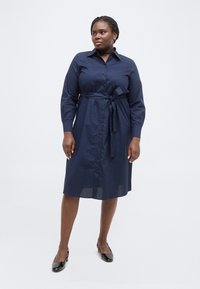 Γυναίκα που στέκεται, φορώντας ένα μπλε φανελένιο φόρεμα μήκους μέχρι το γόνατο με μακριά μανίκια, κουμπιά μπροστά, ζώνη στη μέση και μαύρα ματ λουστρινένια παπούτσια με λουριά.