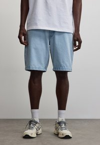Person steht drinnen und trägt ein weißes T-Shirt, hellblaue Jeansshorts, weiße Crew-Socken und weiße Sneakers mit grauen Details.
